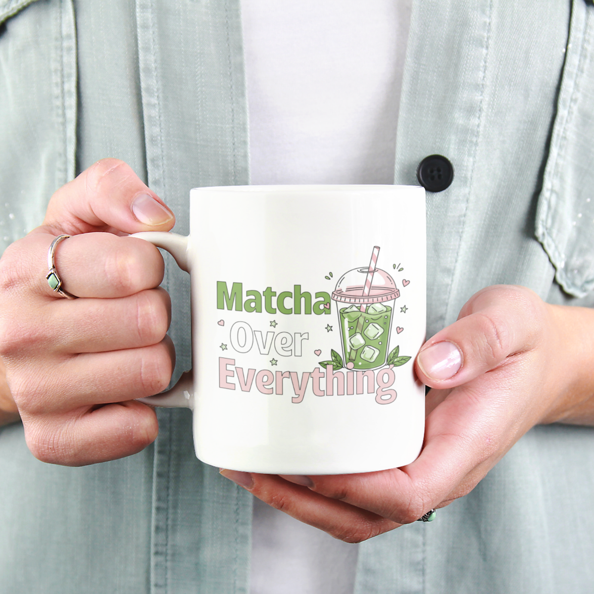Close-up of a person holding a white ceramic matcha mug with an iced matcha latte illustration surrounded by tiny hearts and stars and the text “Matcha Over Everything”