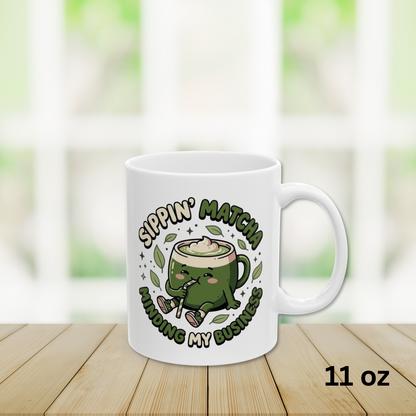 Close-up view 11oz White ceramic matcha mug with a kawaii matcha-cup character and the text “Sippin’ Matcha Minding My Business