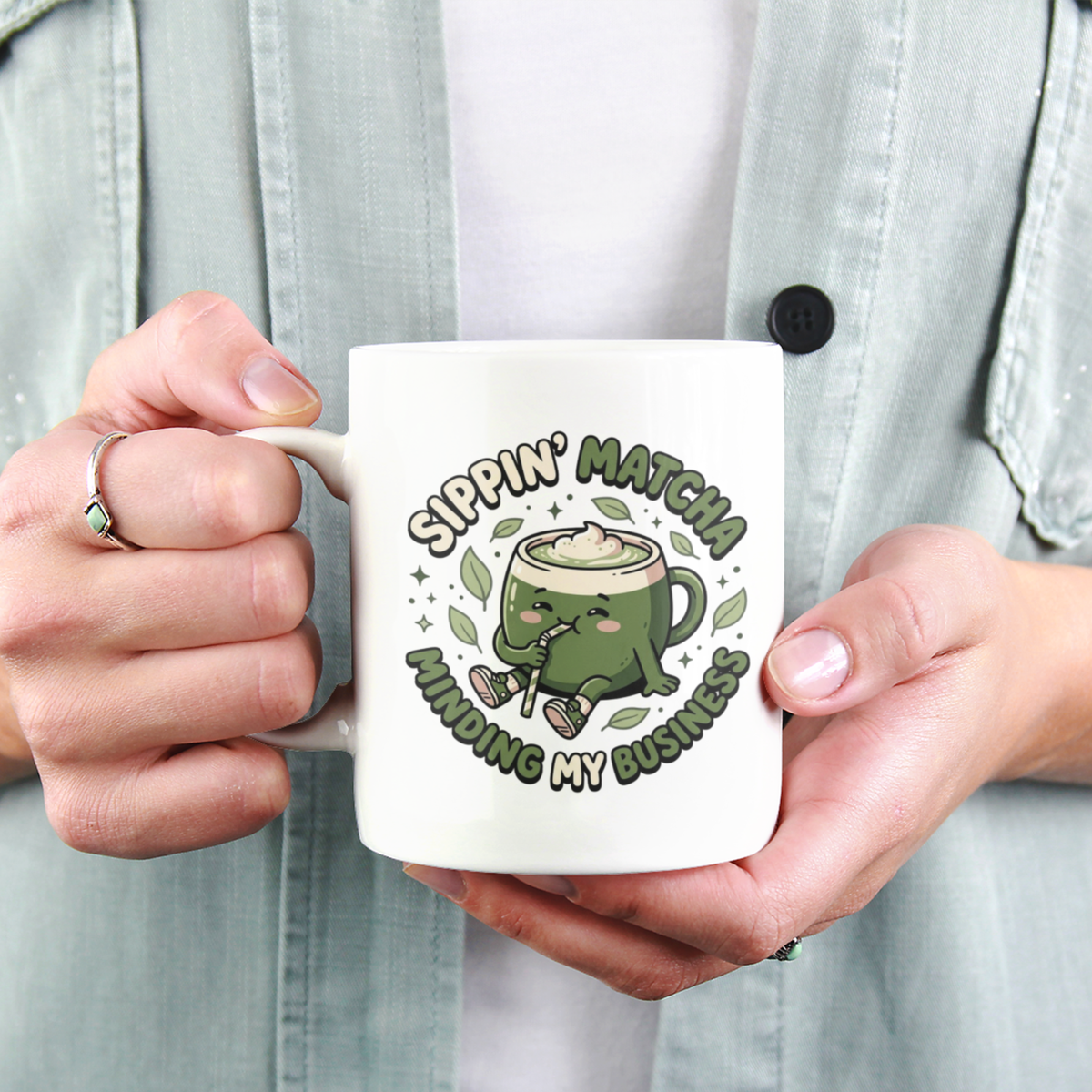 A close-up view of a person holding a white ceramic matcha mug with a kawaii matcha-cup character and the text “Sippin’ Matcha, Minding My Business"