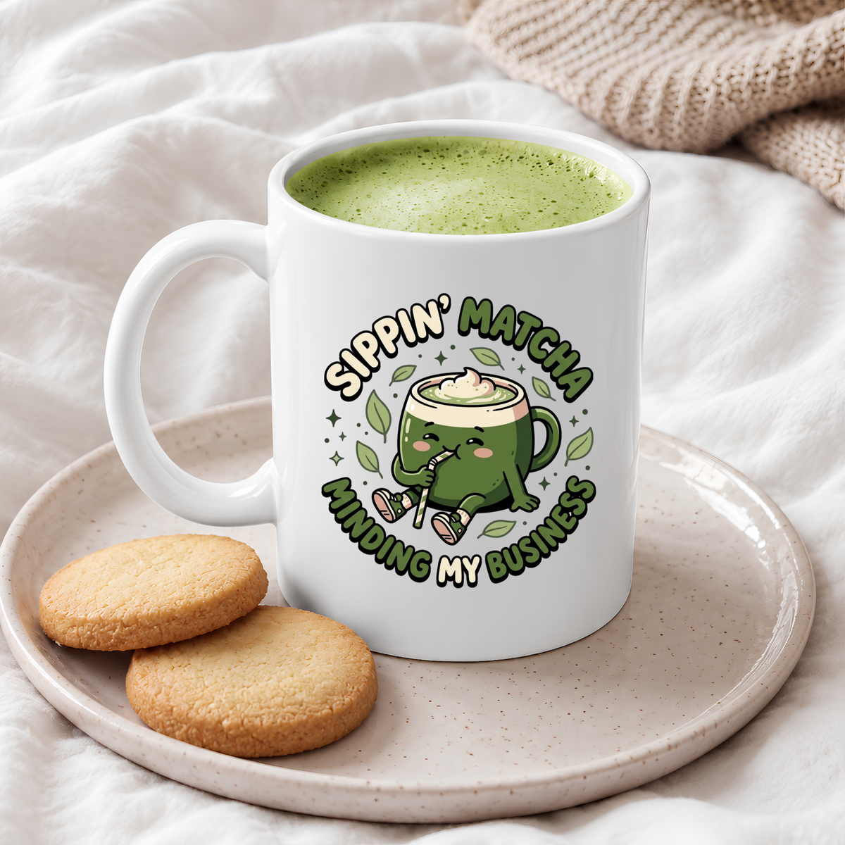 White ceramic matcha mug with a kawaii matcha-cup character and the text “Sippin’ Matcha Minding My Business,” filled with a green matcha latte on a plate with shortbread cookies