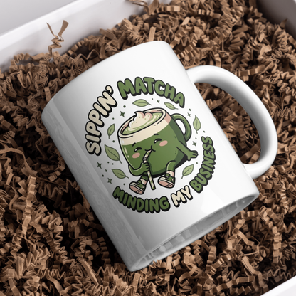 White ceramic matcha mug with a kawaii matcha-cup character and the text “Sippin’ Matcha Minding My Business, resting in brown paper shred in a gift box