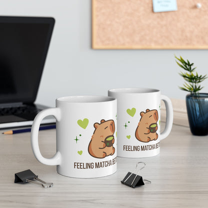 White ceramic matcha mug with a cute capybara drinking green tea, surrounded by hearts and stars, and the text “Feeling Matcha Better,” view of both sides