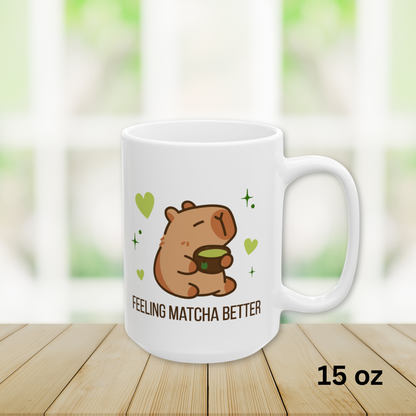 Close-up of a white ceramic matcha mug with a cute capybara drinking green tea, surrounded by hearts and stars, and the text “Feeling Matcha Better,” 15oz