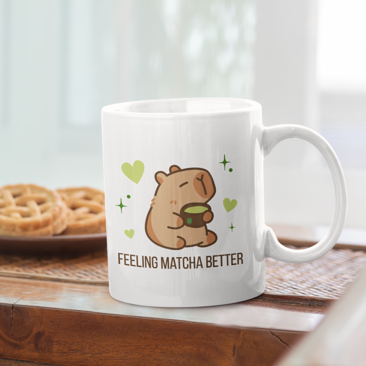 White ceramic matcha mug with a cute capybara drinking green tea, surrounded by hearts and stars, and the text “Feeling Matcha Better,” resting next to a plate of cookies