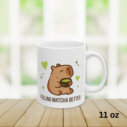 Close-up of a white ceramic matcha mug with a cute capybara drinking green tea, surrounded by hearts and stars, and the text “Feeling Matcha Better,” 11oz