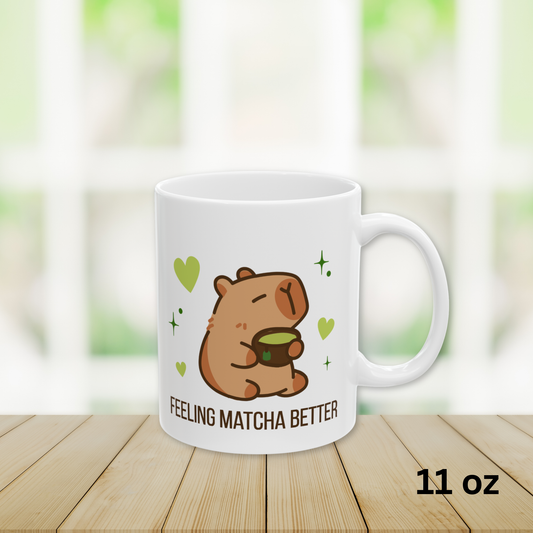 Close-up of a white ceramic matcha mug with a cute capybara drinking green tea, surrounded by hearts and stars, and the text “Feeling Matcha Better,” 11oz
