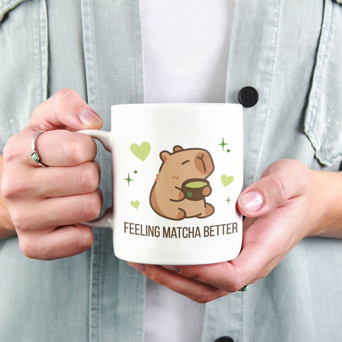 A close-up view of a person holding a white ceramic matcha mug with a cute capybara drinking green tea and the text “Feeling Matcha Better"
