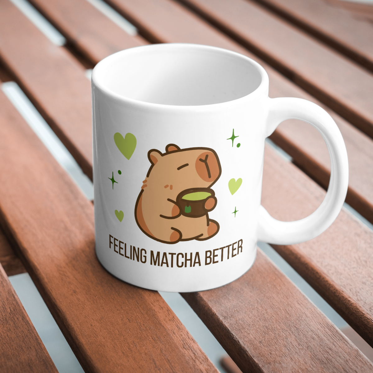 White ceramic matcha mug with a cute capybara drinking green tea, surrounded by hearts and stars, and the text “Feeling Matcha Better,” resting on a wooden table