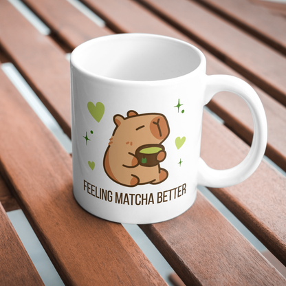 White ceramic matcha mug with a cute capybara drinking green tea, surrounded by hearts and stars, and the text “Feeling Matcha Better,” resting on a wooden table