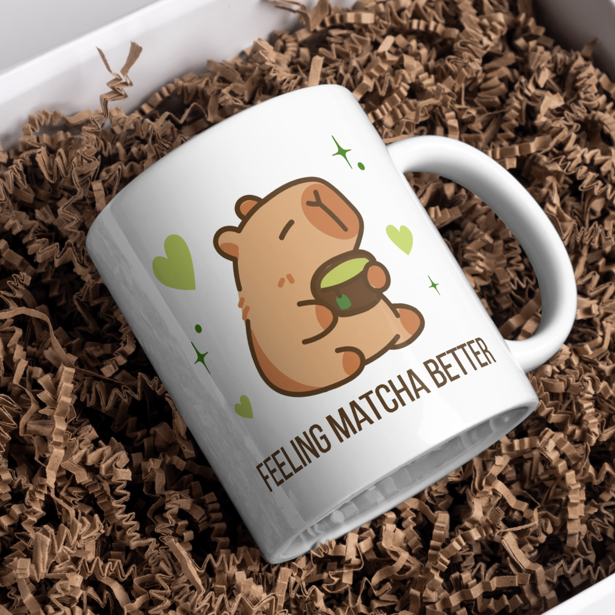 White ceramic matcha mug with a cute capybara drinking green tea, surrounded by hearts and stars, and the text “Feeling Matcha Better,” resting in brown paper shreds in a gift box