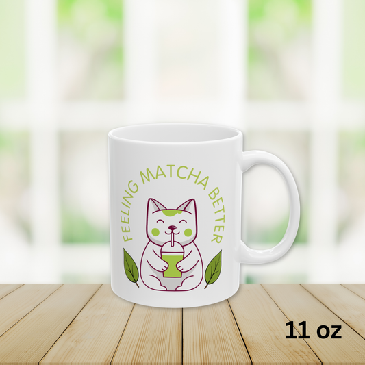 Close-up view of a white ceramic matcha mug with a cute cat drinking matcha and the text “Feeling Matcha Better," 11oz