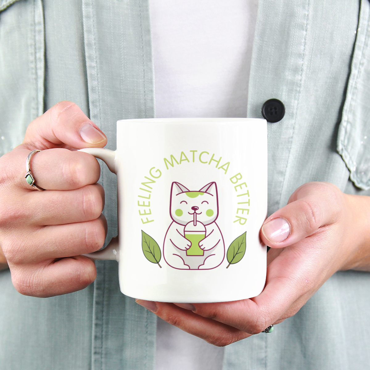 A close-up view of a person holding a white ceramic matcha mug with a cute cat drinking matcha and the text “Feeling Matcha Better”