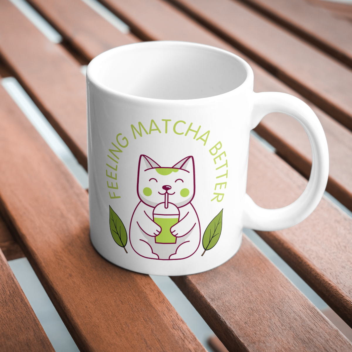 White ceramic matcha mug with a cute cat drinking matcha and the text “Feeling Matcha Better,” resting on a wooden table