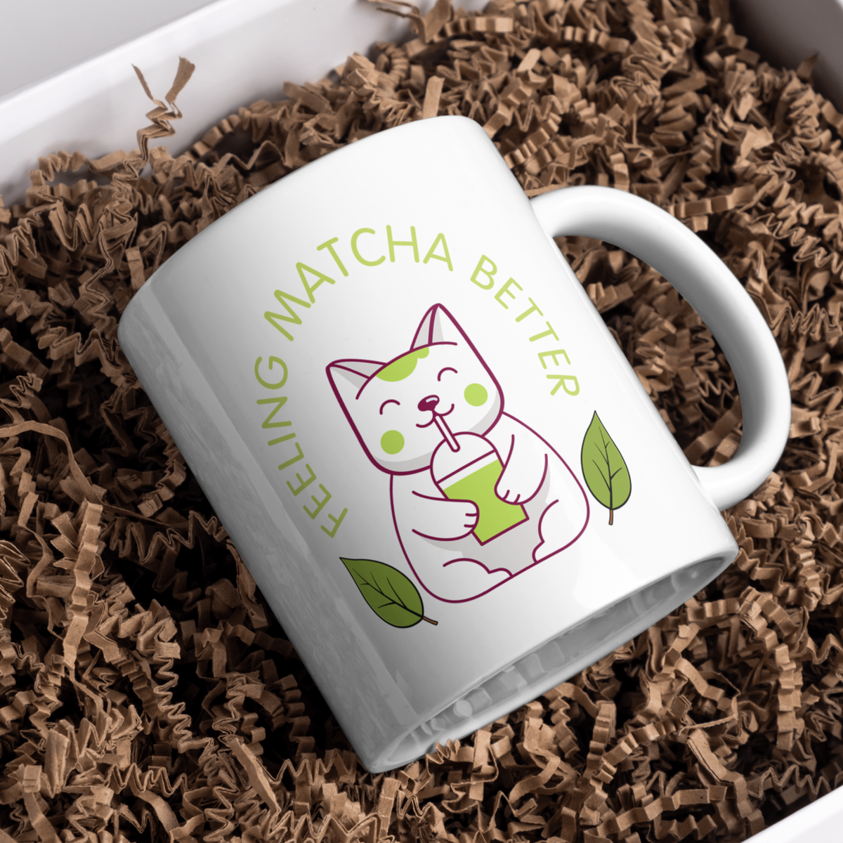 White ceramic matcha mug with a cute cat drinking matcha and the text “Feeling Matcha Better,” resting in brown shreds in a gift box