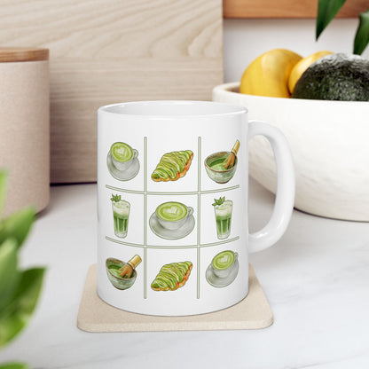 White ceramic mug with a matcha tic-tac-toe design of green lattes, matcha croissants, iced matcha, and whisked matcha bowls, resting on a coaster on a kitchen counter