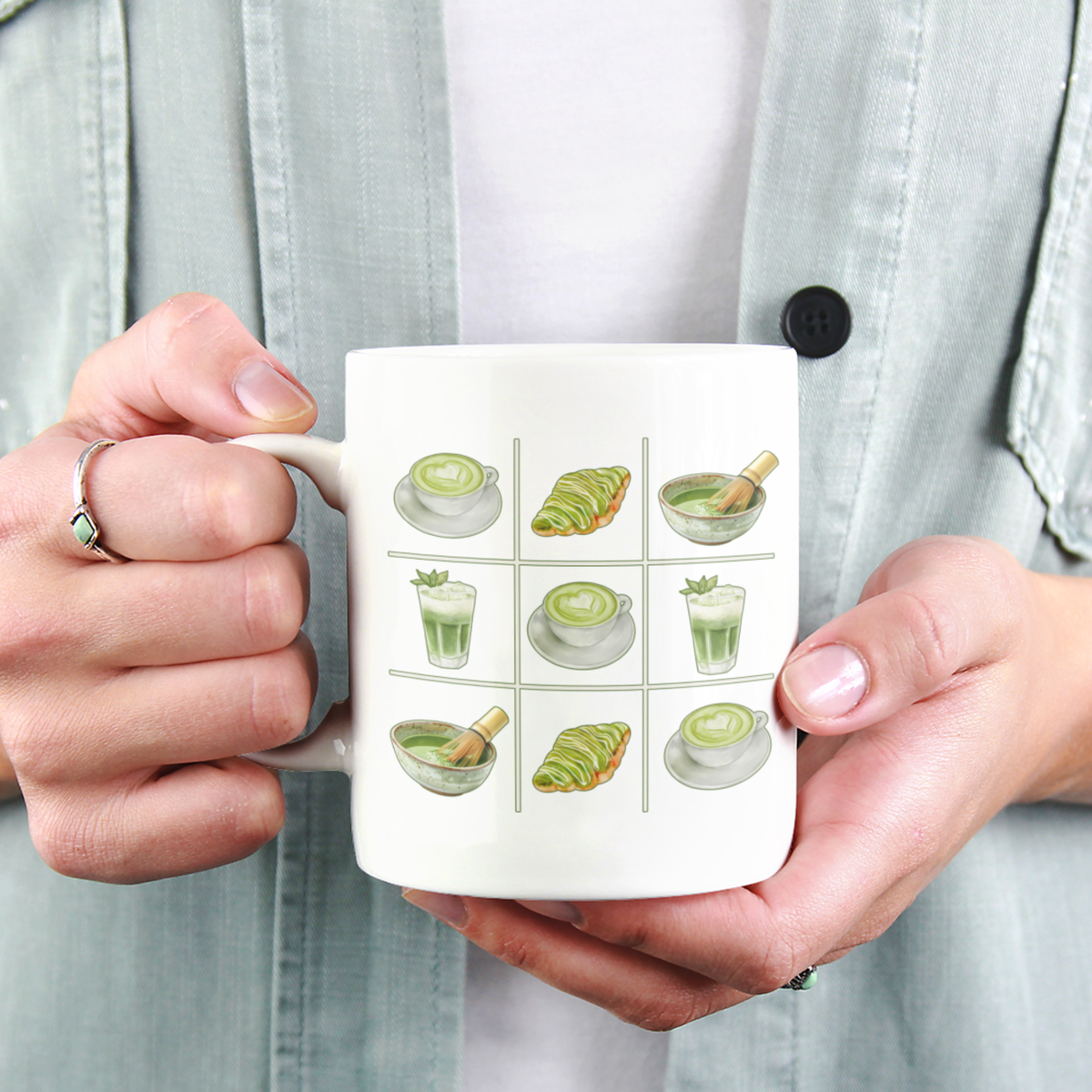 Close-up view of a person holding a white ceramic mug with a matcha tic-tac-toe design of green lattes, matcha croissants, iced matcha, and whisked matcha bowls