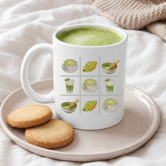 White ceramic mug with a matcha tic-tac-toe design of green lattes, matcha croissants, iced matcha, and whisked matcha bowls, filled with a green matcha latte on a plate with shortbread cookies