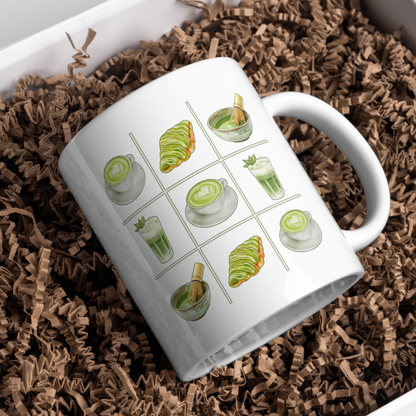White ceramic mug with a matcha tic-tac-toe design of green lattes, matcha croissants, iced matcha, and whisked matcha bowls, resting in brown paper shred in a gift box