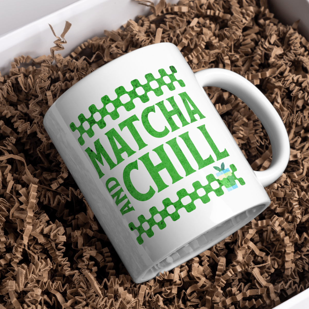 
White ceramic matcha mug with a checkered border above and below the text “Matcha and Chill,” resting in brown paper shreds in a gift box
