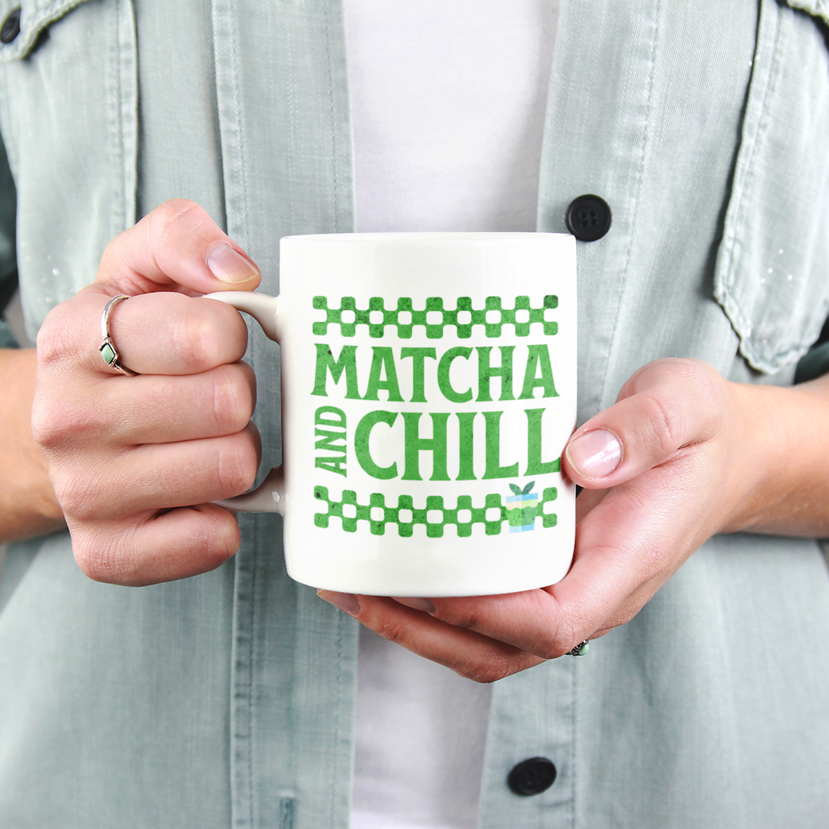 Close-up of a person holding a white ceramic matcha mug with a checkered border above and below the text “Matcha and Chill" 