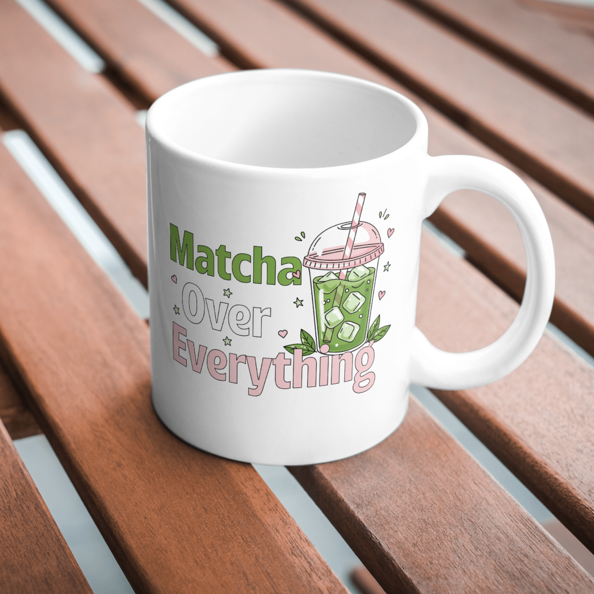 White ceramic matcha mug with an iced matcha latte illustration surrounded by tiny hearts and stars and the text “Matcha Over Everything,” resting on a wooden table