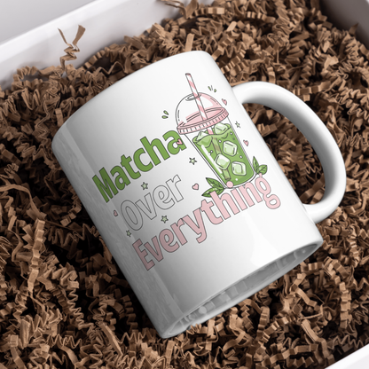 White ceramic matcha mug with an iced matcha latte illustration surrounded by tiny hearts and stars and the text “Matcha Over Everything,” resting in brown paper shreds in a gift box