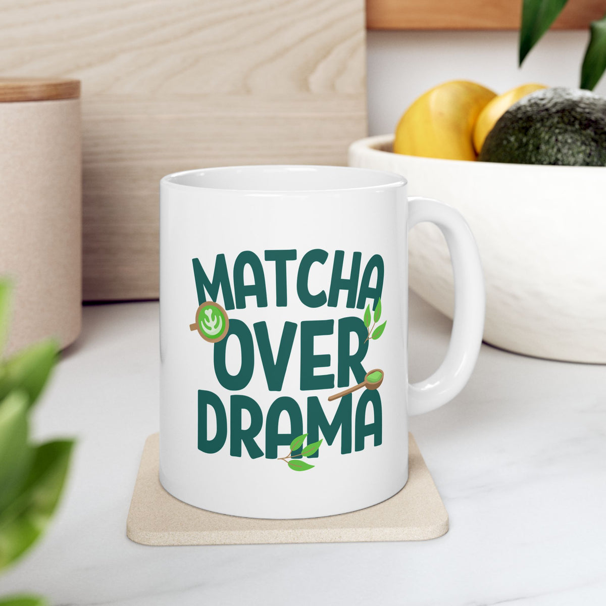 White ceramic "Matcha Over Drama" coffee mug with mini images of green tea leaves, a matcha latte, and wooden spoon filled with matcha powder, sitting on a kitchen counter