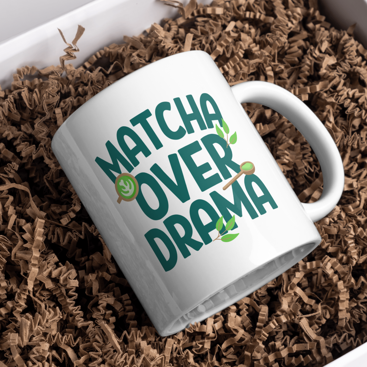 White ceramic "Matcha Over Drama" coffee mug with mini images of green tea leaves, a matcha latte, resting in brown paper shred in a gift box