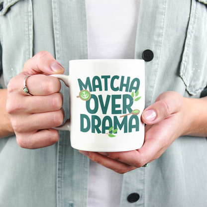 A close-up view of a person holding a white ceramic "Matcha Over Drama" coffee mug with mini images of green tea leaves, a matcha latte, and a wooden spoon filled with matcha powder