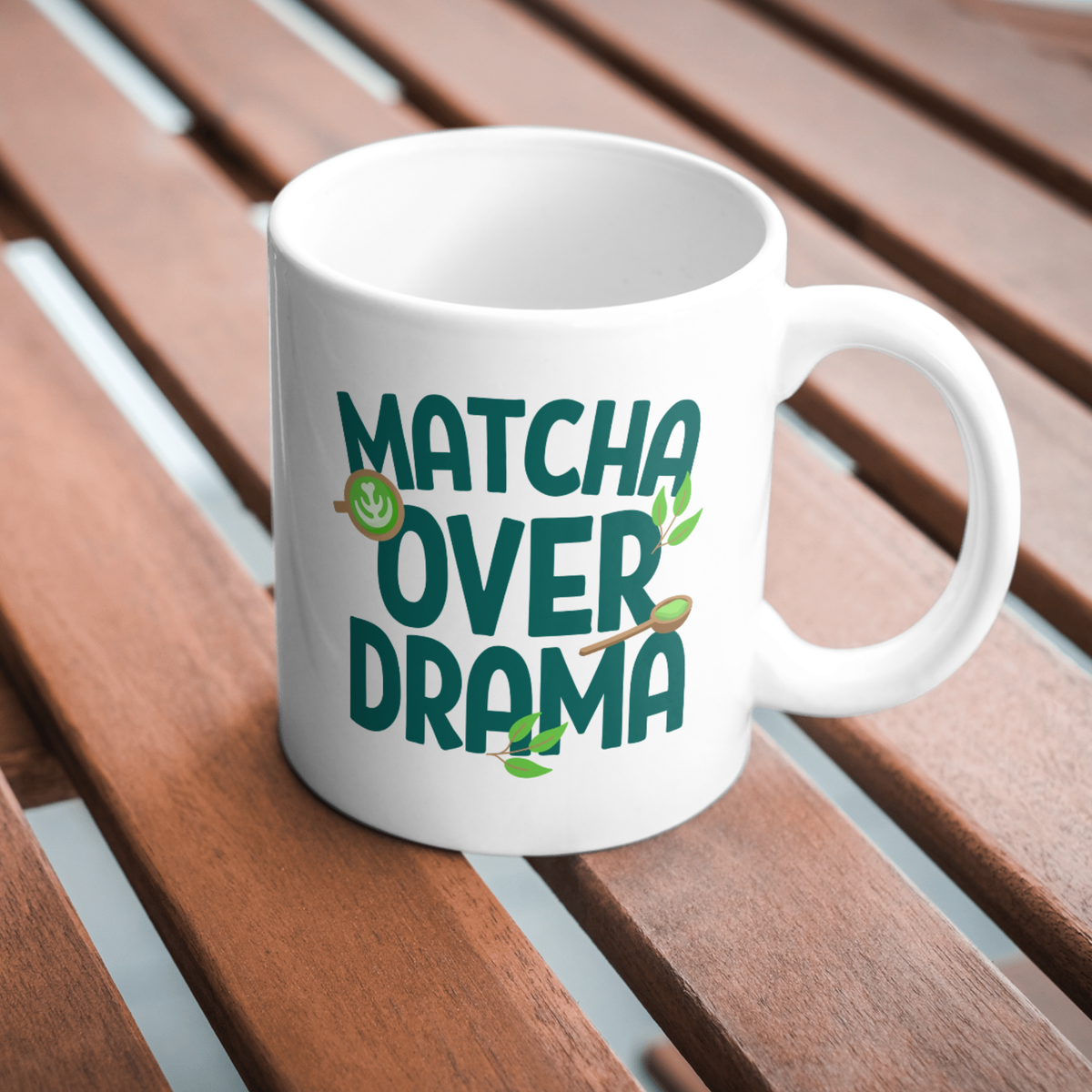 White ceramic "Matcha Over Drama" coffee mug with mini images of green tea leaves, a matcha latte, and wooden spoon filled with matcha powder, sitting a wooden table