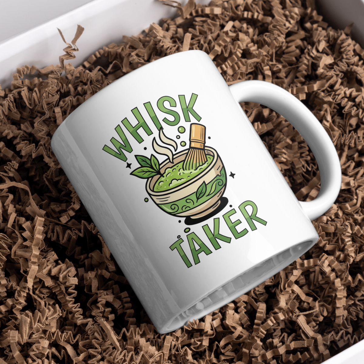 White ceramic mug with the text “Whisk Taker” and an illustration of a steaming matcha bowl with a bamboo whisk and tea leaves, resting in brown paper shred in a gift box