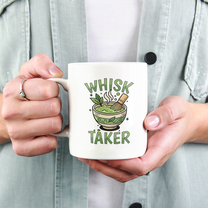 A close-up view of a person holding a white ceramic mug with the text “Whisk Taker” and an illustration of a steaming matcha bowl with a bamboo whisk and tea leaves