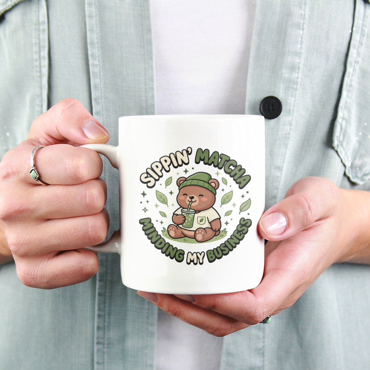 A close-up view of a person holding a white ceramic matcha mug with a bear drinking an iced matcha and the text “Sippin’ Matcha, Minding My Business"